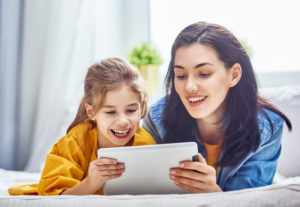 Happy loving family. Young mother and her daughter girl are playing in bedroom. Funny mom and lovely child are having fun with tablet.